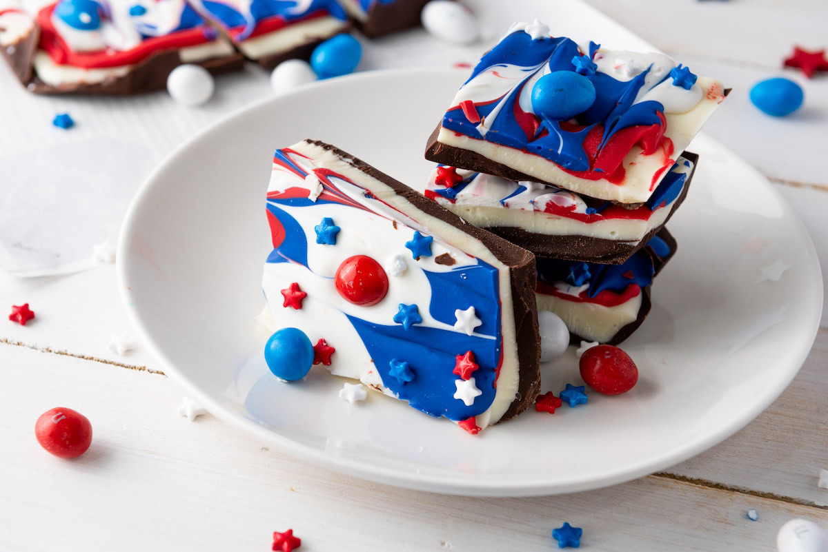 A plate with stacked pieces of red, white, and blue swirled chocolate bark, decorated with candy stars and coated candies, on a white wooden surface.