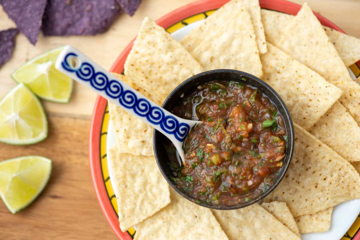A bowl of salsa with a spoon is surrounded by tortilla chips on a plate, with lime wedges and blue corn chips on the side.