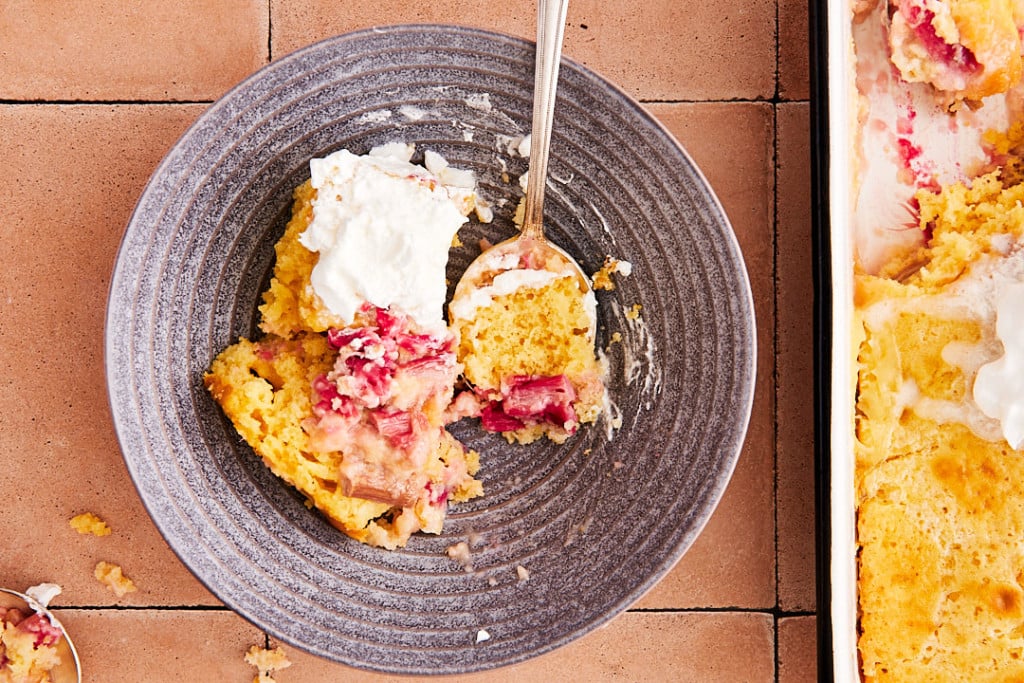 A serving of fruit cobbler with whipped cream on a gray plate, partially eaten with a spoon, next to a baking dish of cobbler on a tiled surface.