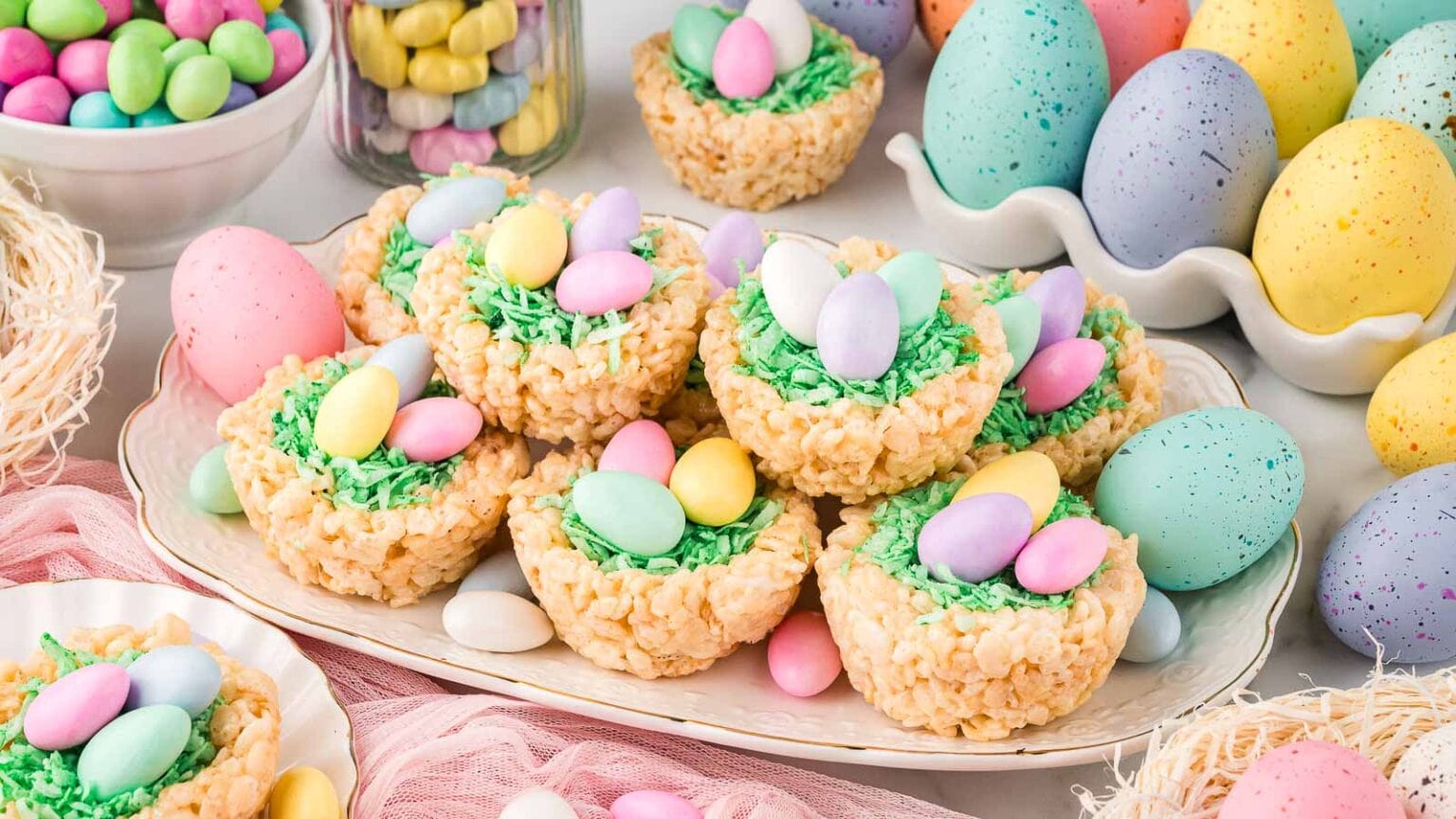 A plate of Rice Krispies treat nests filled with green coconut shreds and pastel candy eggs, surrounded by colorful candy eggs and decorative Easter eggs.