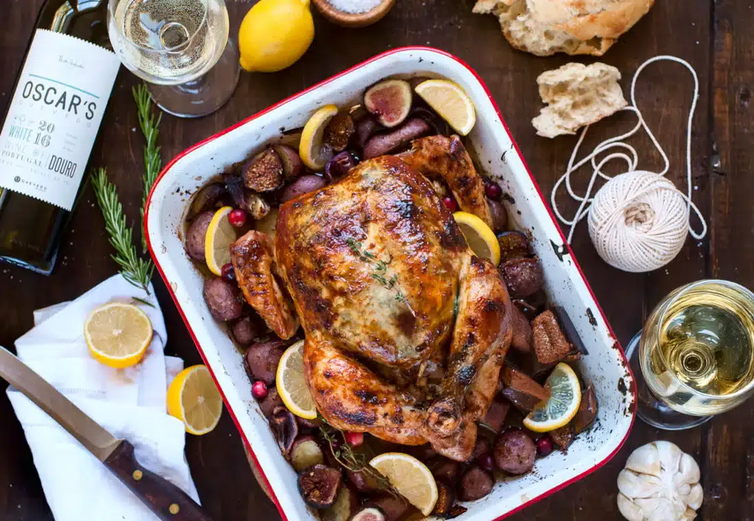 Roast chicken with lemons, figs, and potatoes in a pan, surrounded by a wine bottle, wine glasses, bread, string, and a knife on a wooden table.