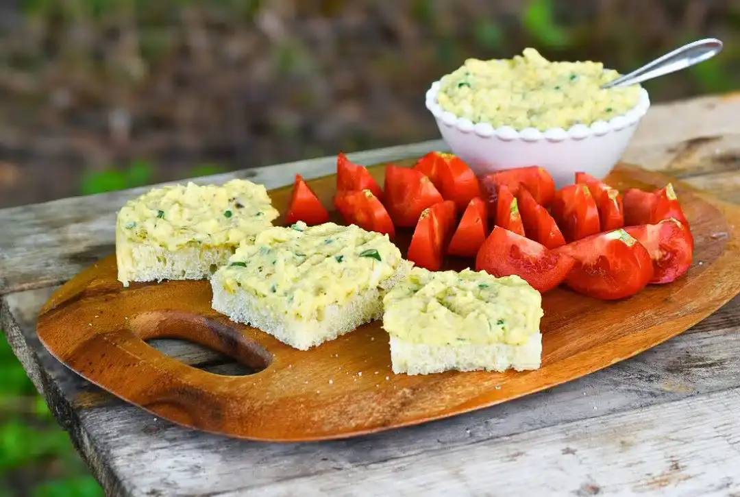 A wooden board with slices of bread topped with a creamy spread, tomato wedges, and a bowl of the same spread with a spoon, set on a rustic outdoor table.