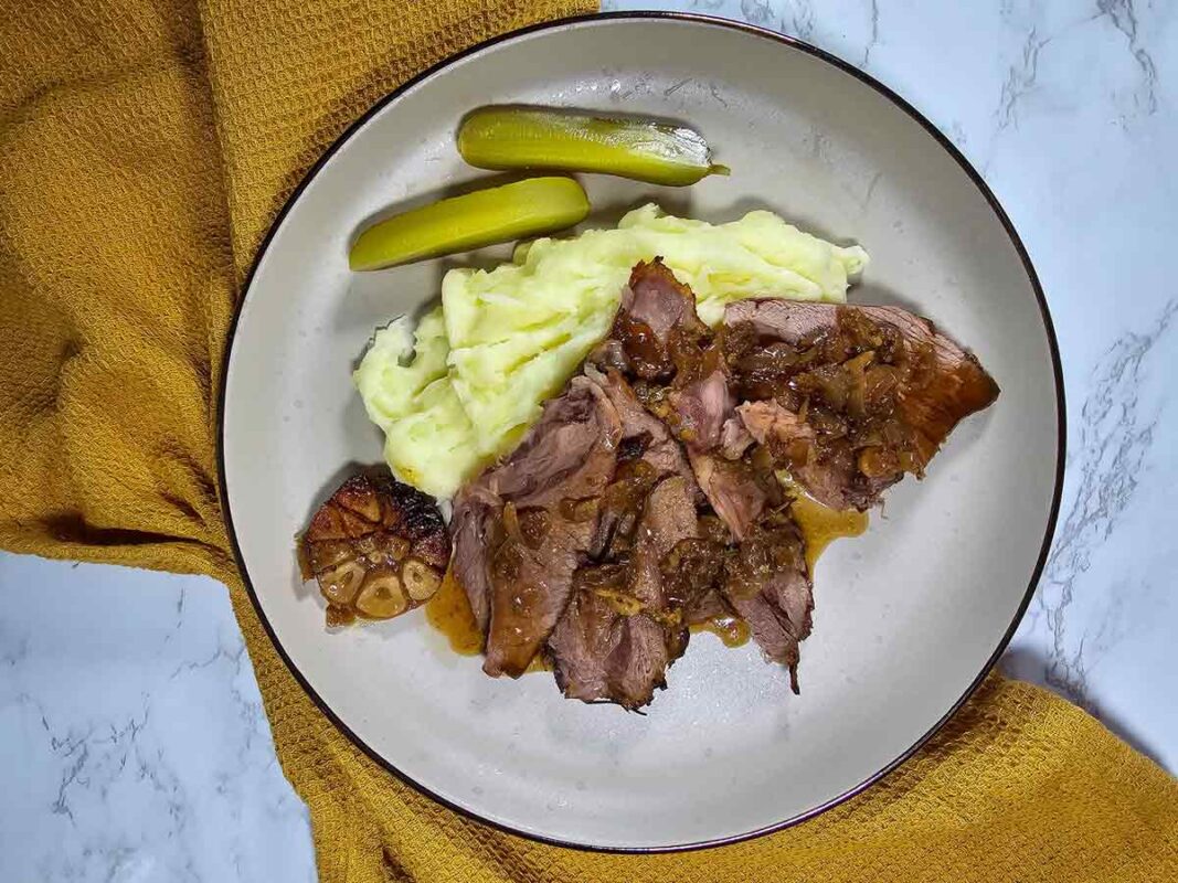 Plate with sliced roast meat topped with onions, mashed potatoes, two pickles, and a head of roasted garlic on a white dish, set on a yellow textured cloth.