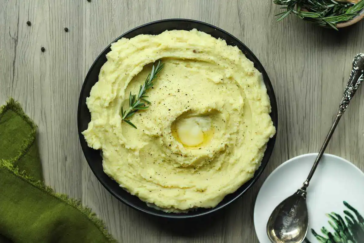 A bowl of mashed potatoes topped with a pat of melting butter and a sprig of rosemary, placed on a wooden table with a spoon and green cloth nearby.