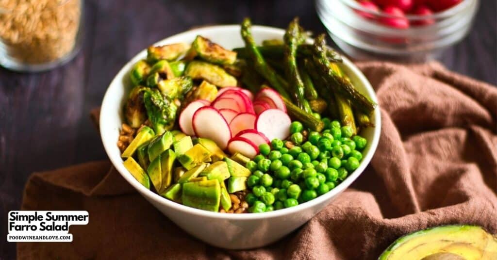 A bowl of farro salad topped with sliced avocado, peas, radishes, roasted asparagus, and zucchini sits on a brown cloth, with a partial view of avocado and radishes nearby.