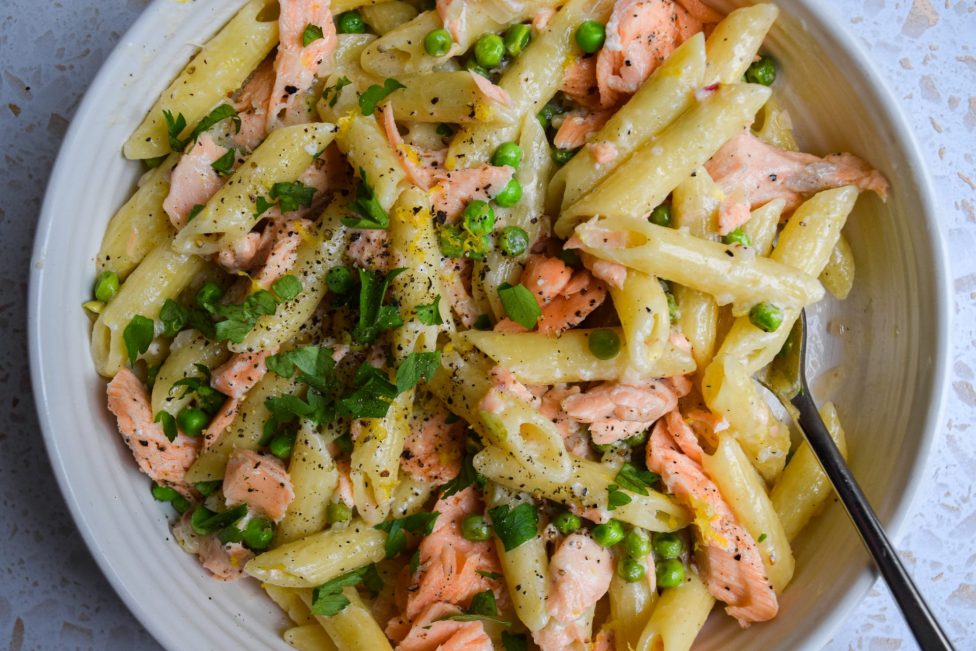 A bowl of penne pasta with pieces of salmon, green peas, chopped herbs, and black pepper, served with a creamy sauce.
