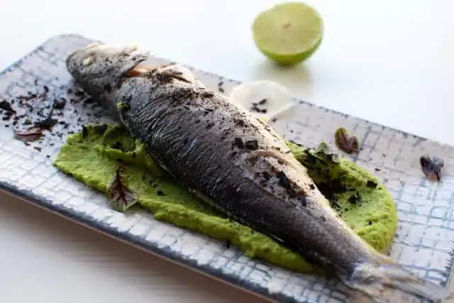Whole cooked fish served on a rectangular plate over a bed of green puree, garnished with seasoning, with a lime half in the background.