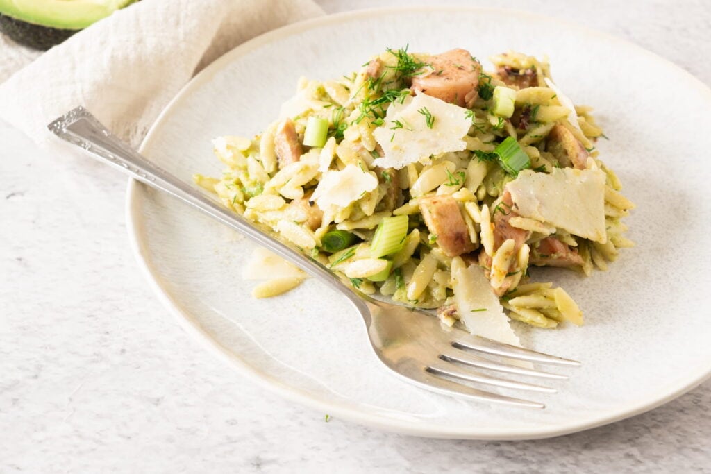 A plate of orzo pasta salad with pieces of grilled chicken, shaved parmesan, and green herbs, served with a fork on a light-colored plate.
