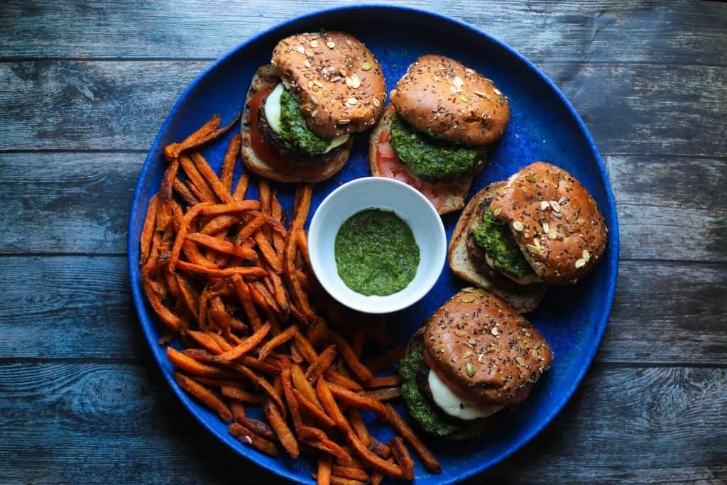 A blue plate with three veggie burgers topped with green sauce and tomato, sweet potato fries on the side, and a bowl of green dipping sauce in the center.