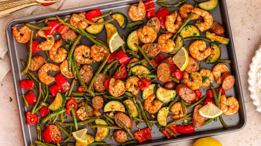 Sheet pan with cooked shrimp, sausage slices, green beans, zucchini, red bell peppers, and lemon wedges arranged evenly.