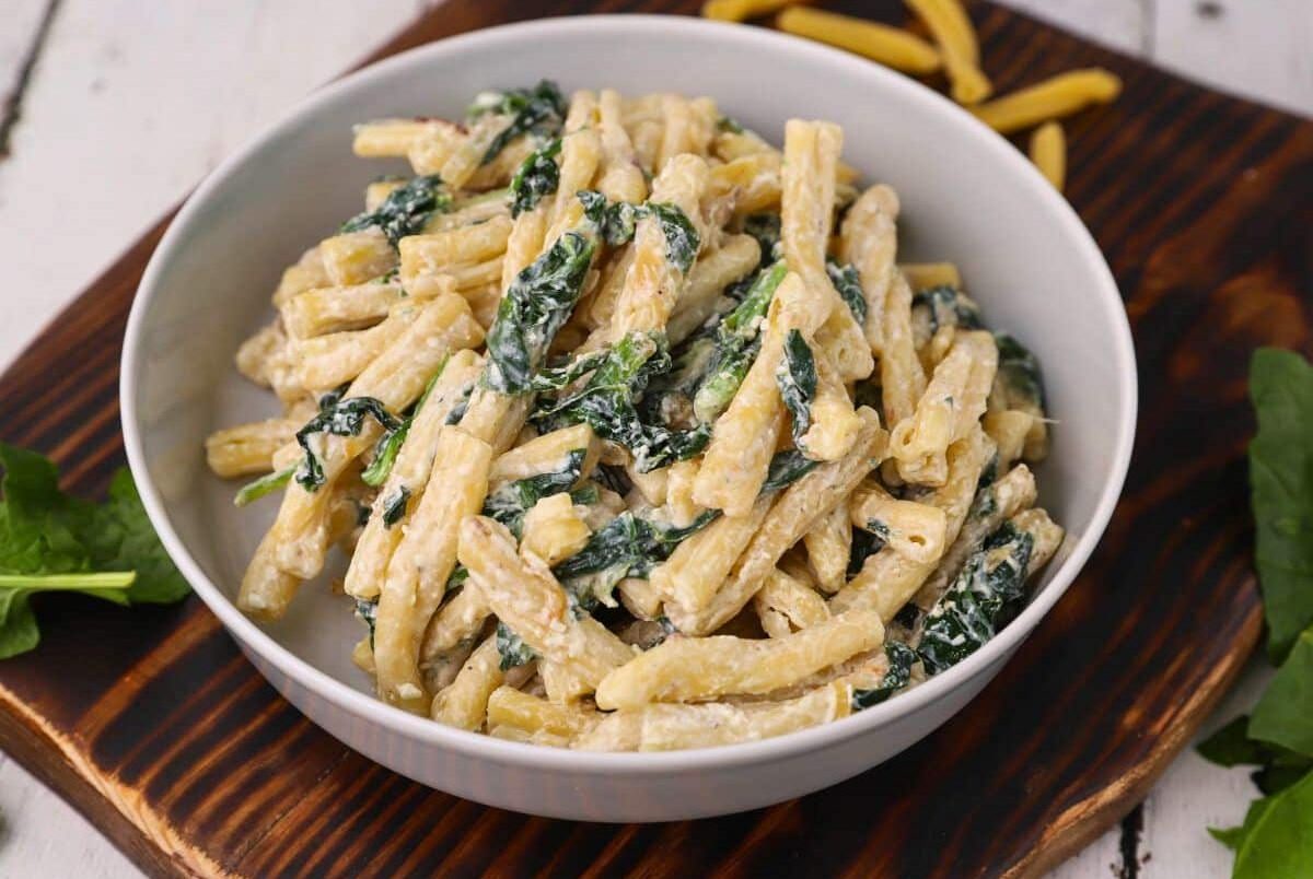 A bowl of creamy pasta with spinach, served on a wooden board with some uncooked pasta and spinach leaves nearby.