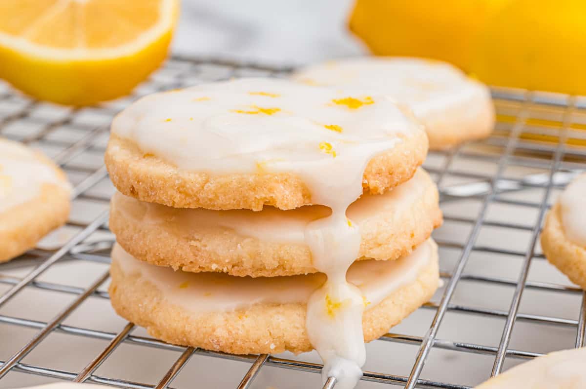 Three stacked lemon cookies with icing dripping down, placed on a metal cooling rack, with a lemon half in the background.