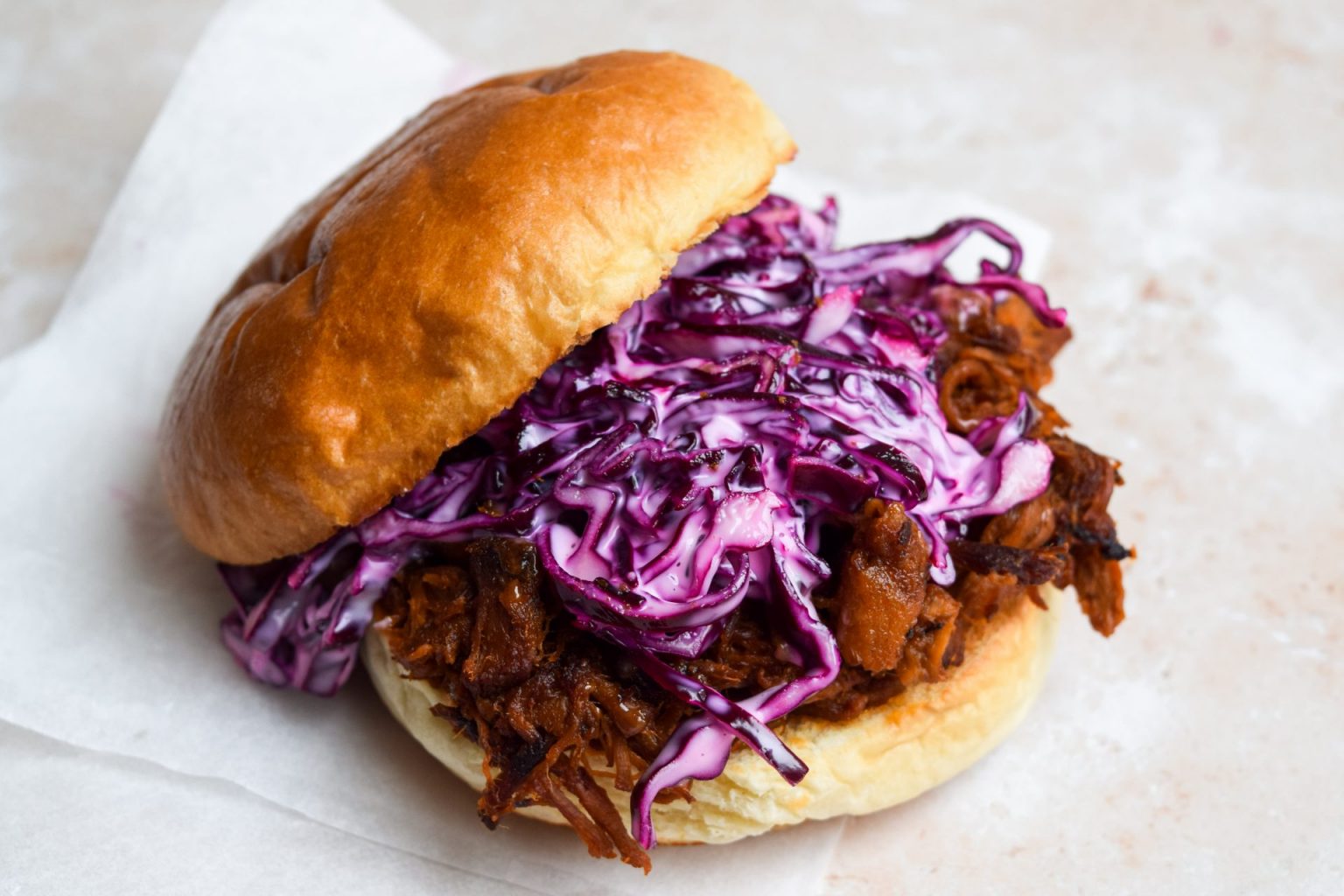 A pulled pork sandwich on a bun topped with creamy purple coleslaw, resting on a piece of white parchment paper.