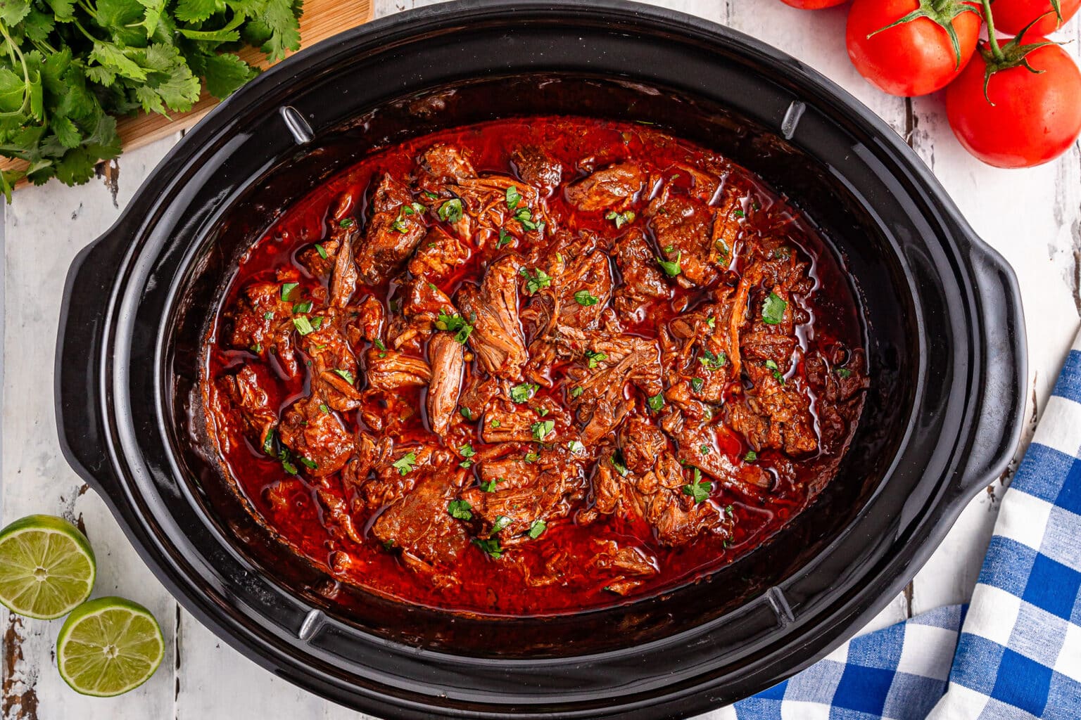 Shredded beef in a rich red sauce inside a slow cooker, garnished with herbs, with tomatoes, cilantro, limes, and a blue checked cloth nearby.