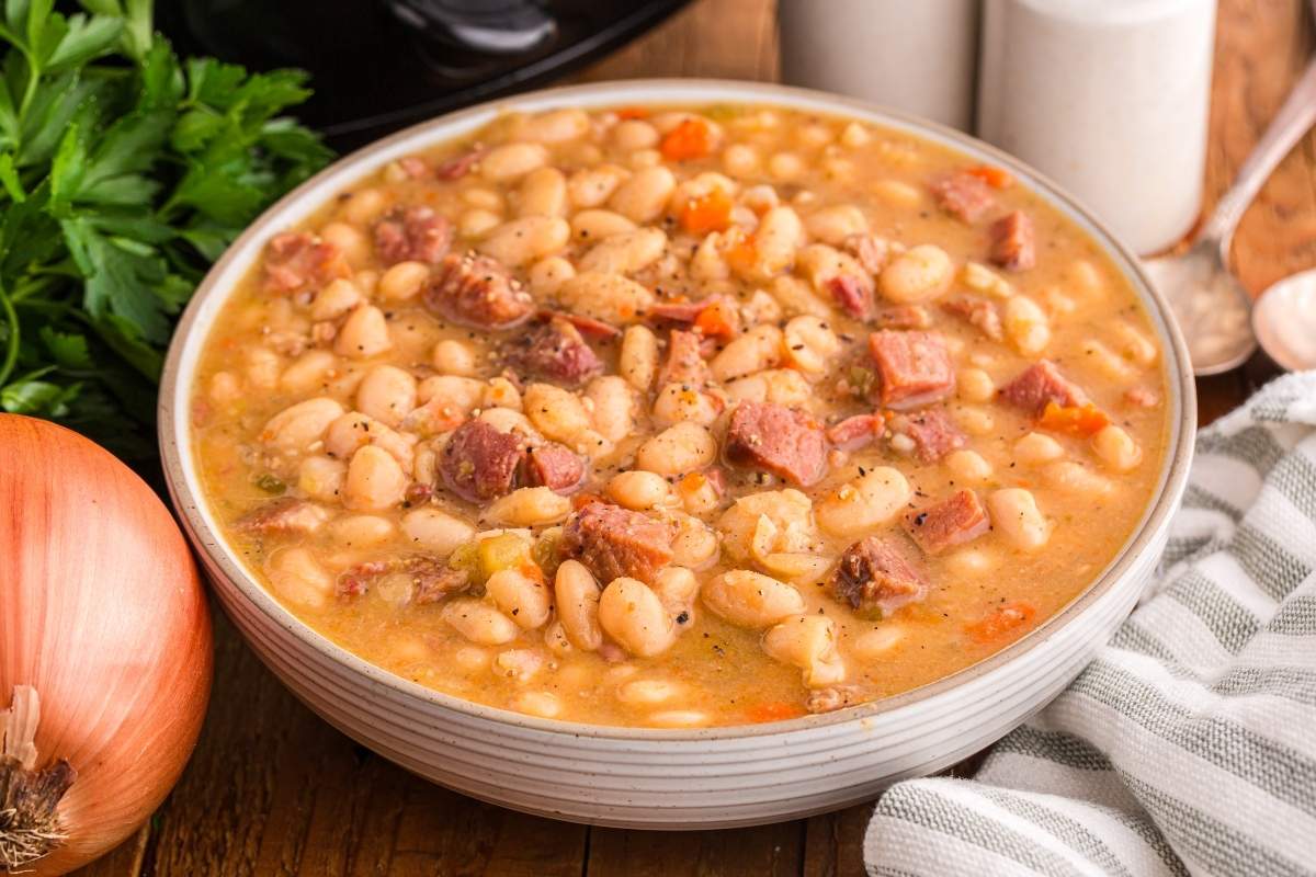 A bowl filled with white bean and ham soup, with visible chunks of ham and carrots, sits on a wooden table next to an onion, parsley, and salt and pepper shakers.