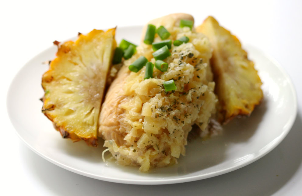 A white plate with baked chicken breast topped with diced green onions and cooked pineapple, garnished with two pineapple wedges.