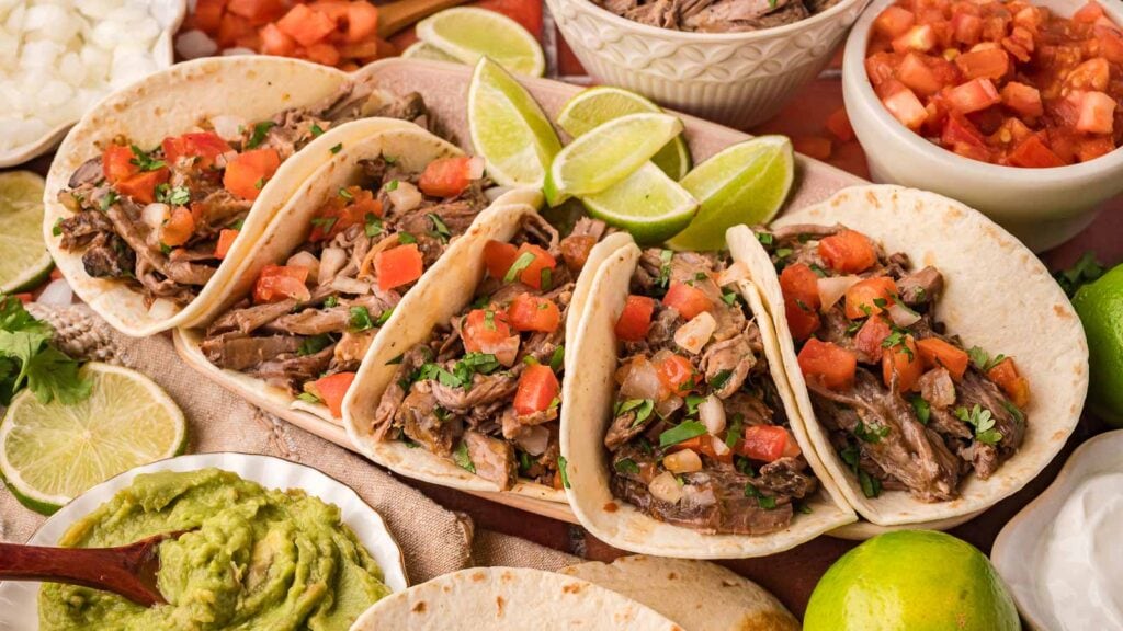 Four beef tacos topped with diced tomatoes and onions are displayed on a platter, surrounded by lime wedges, guacamole, and bowls of salsa.