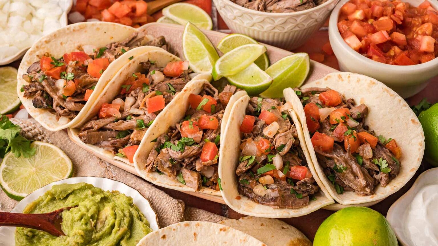 Four beef tacos topped with diced tomatoes, onions, and cilantro are arranged on a platter with lime wedges, guacamole, and salsa on the side.
