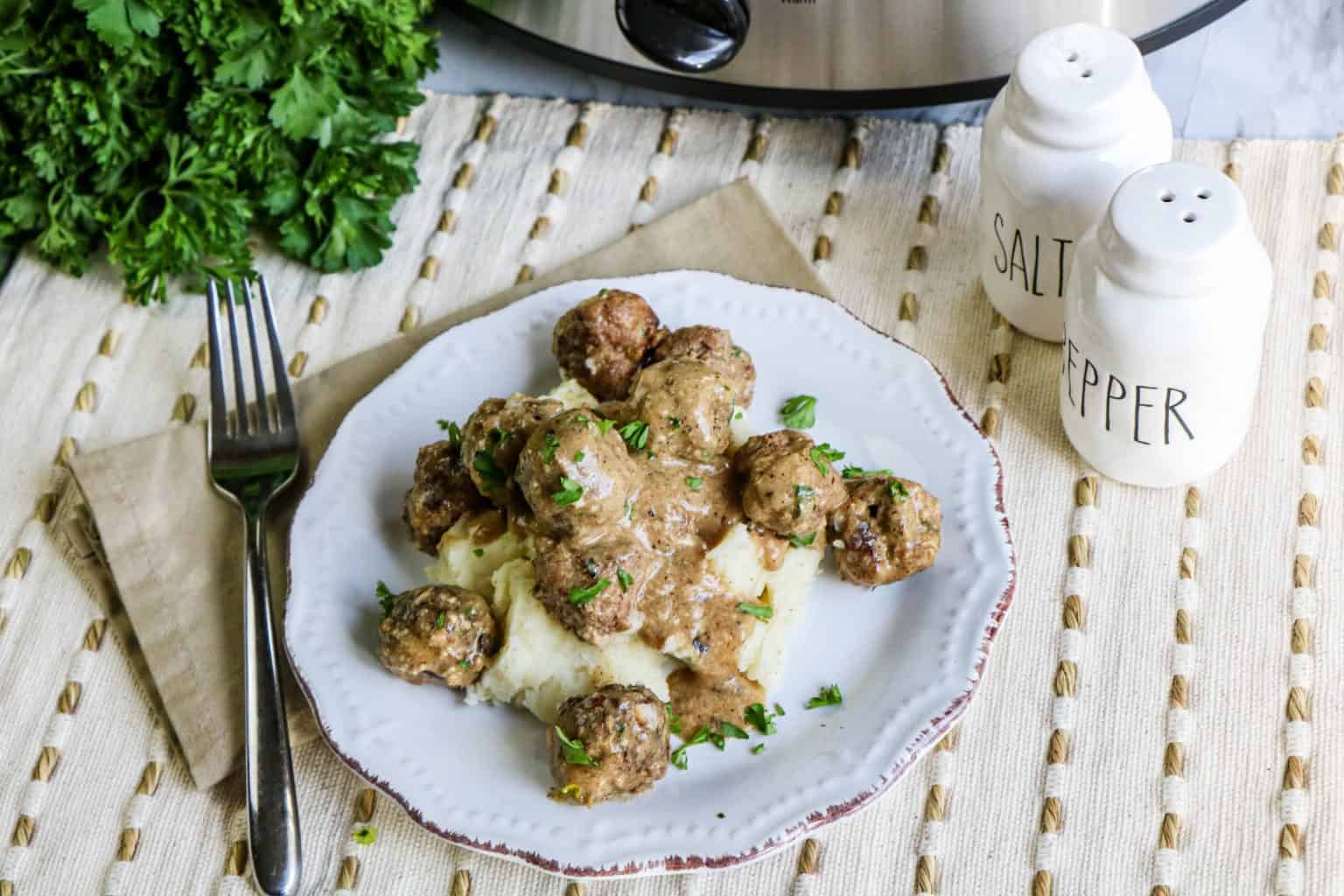 A plate of meatballs with creamy sauce served over mashed potatoes, garnished with herbs, next to a fork, napkin, parsley, and salt and pepper shakers.