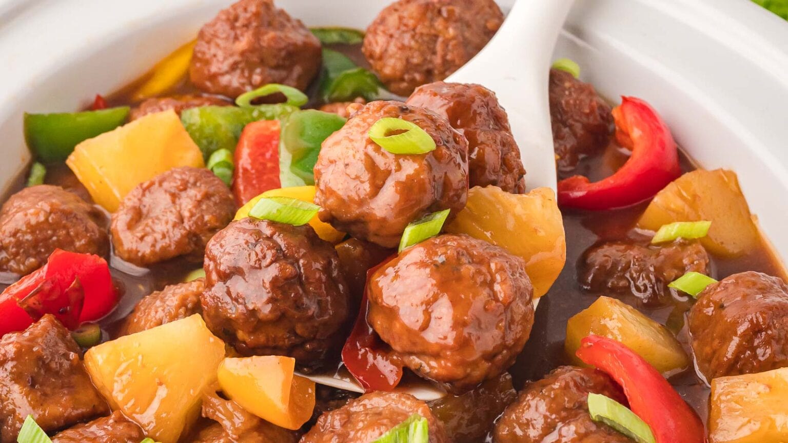 Meatballs in a brown sauce with chunks of pineapple, green onions, and red and green bell peppers, served in a white dish with a serving spoon.
