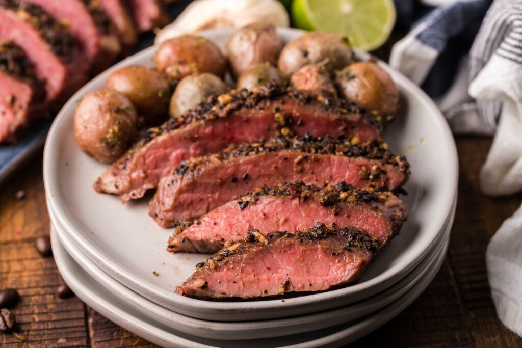 Sliced medium-rare steak with a pepper crust, served with roasted baby potatoes on a white plate.
