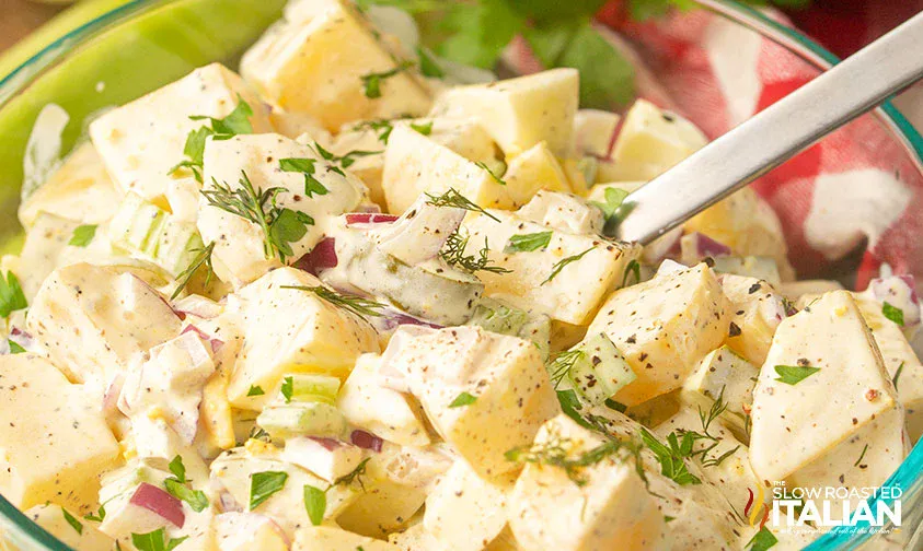 A bowl of creamy potato salad with chopped red onions, celery, herbs, and a pepper seasoning, with a spoon inside.
