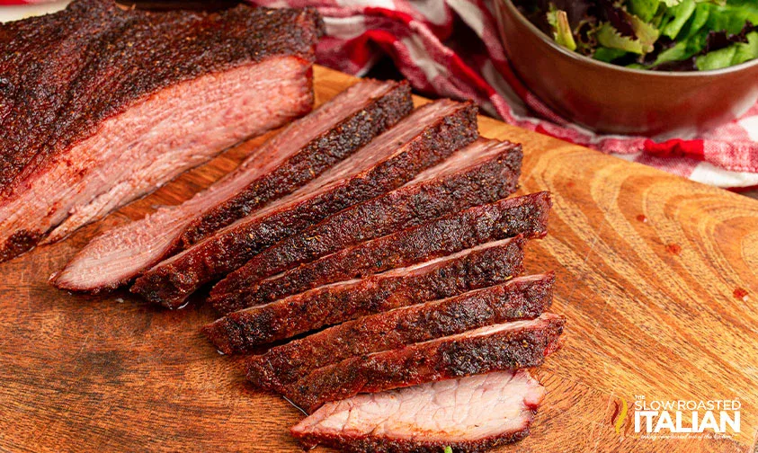 Sliced smoked brisket with a dark seasoned crust on a wooden cutting board, with a bowl of mixed greens and a red-and-white cloth nearby.