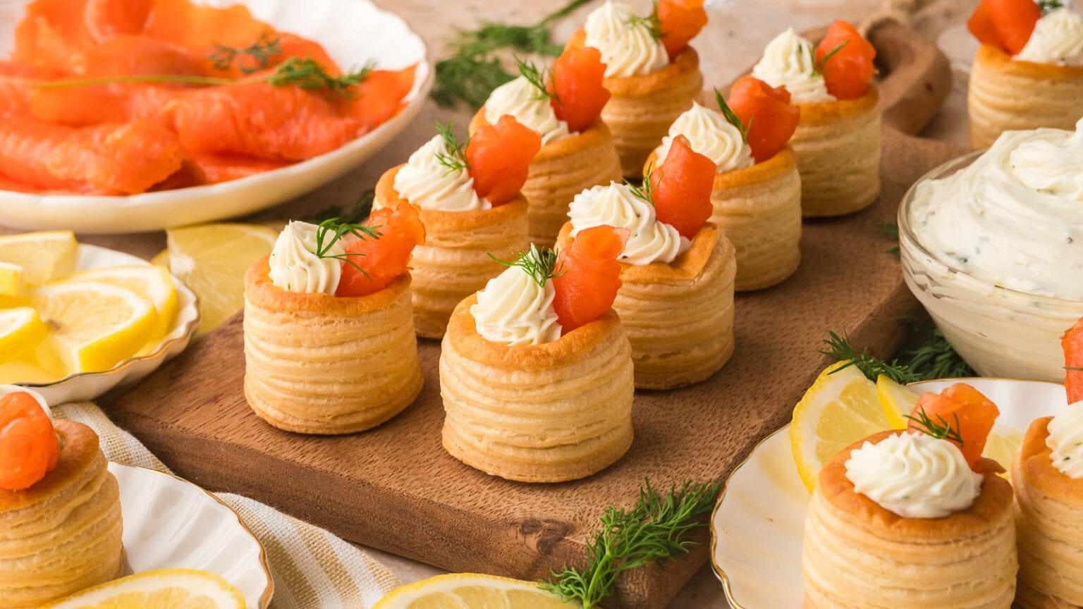 Vol-au-vents filled with cream and topped with smoked salmon and dill are arranged on a wooden board, with plates of lemon slices, smoked salmon, and a bowl of cream cheese nearby.