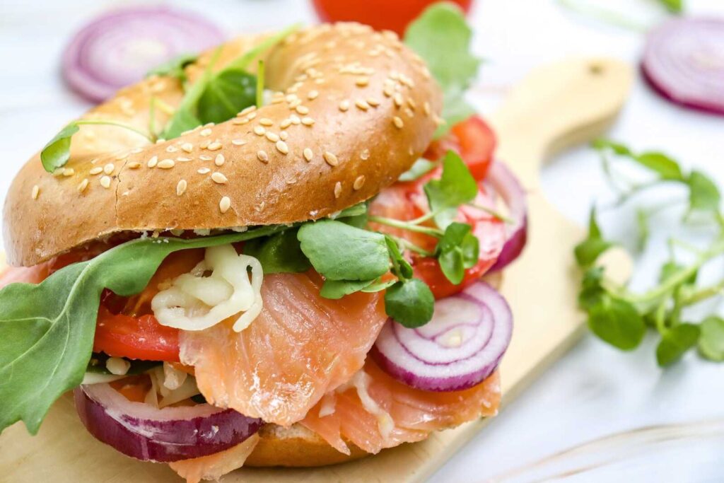 A sesame bagel sandwich filled with smoked salmon, sliced tomato, red onion, arugula, and capers on a wooden board.