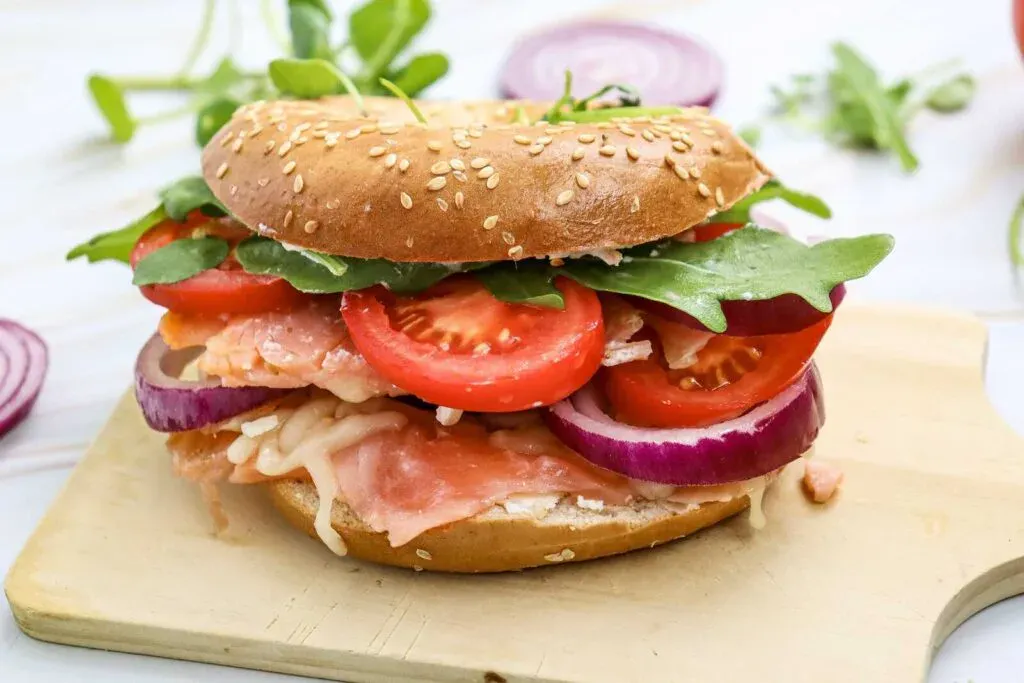 A sesame seed bagel sandwich with smoked salmon, sliced tomatoes, red onion, arugula, and cheese, served on a wooden board.