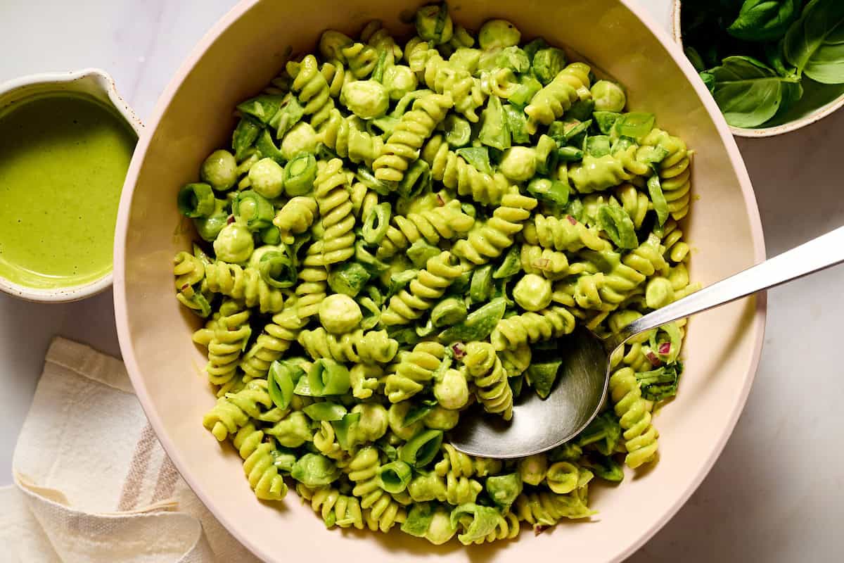 A bowl of rotini pasta mixed with green pesto sauce, mozzarella balls, and spinach, with a serving spoon and a small bowl of extra sauce nearby.