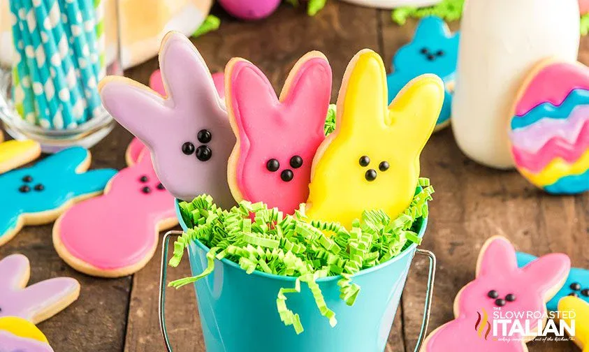 Three bunny-shaped frosted cookies in pastel colors are arranged in a small blue pail with green paper grass. Other decorated cookies and Easter-themed items are in the background.