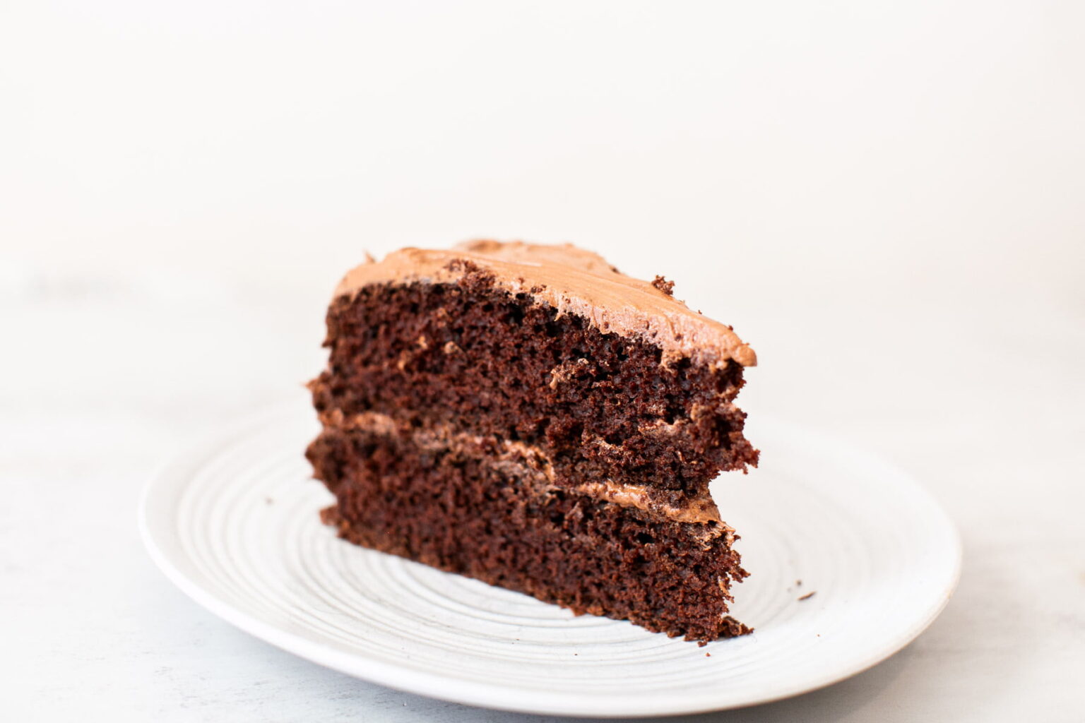 A slice of chocolate layer cake with chocolate frosting on a white plate against a light background.