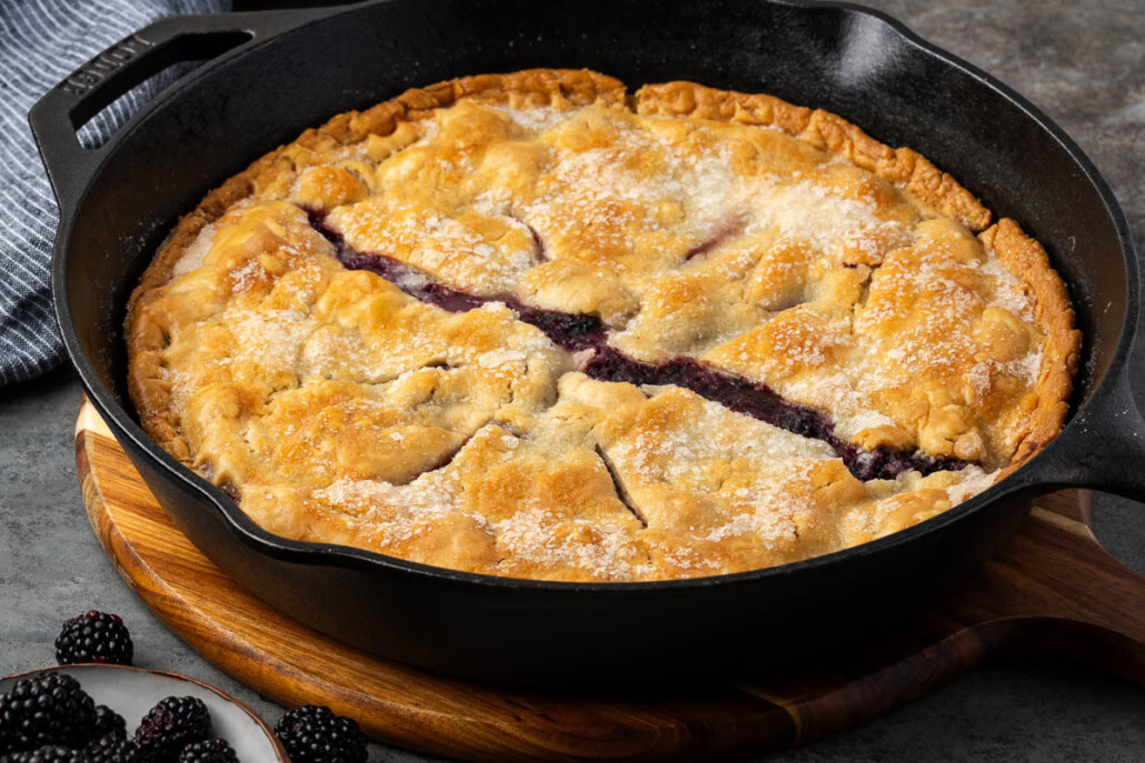 A baked blackberry cobbler with a golden crust is in a black cast iron skillet, with fresh blackberries in a small bowl beside it.