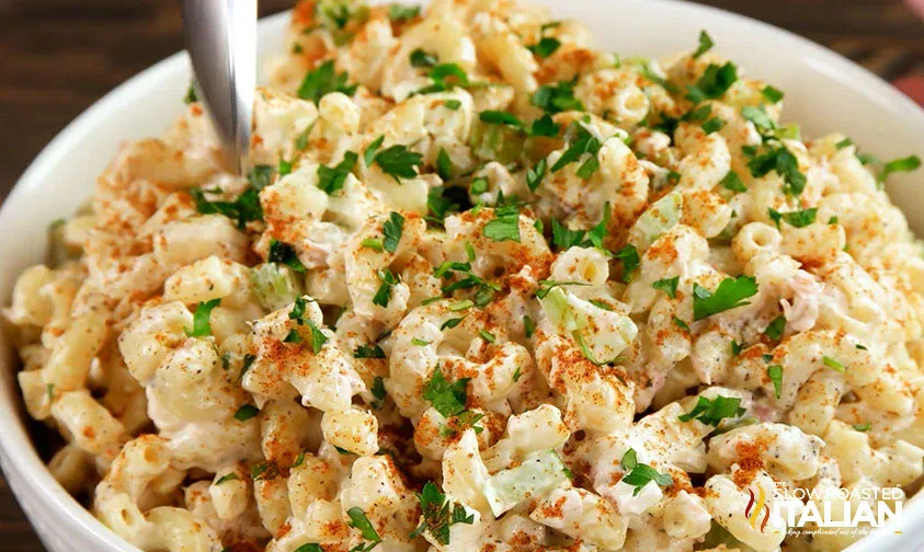 A bowl of creamy macaroni salad topped with chopped parsley and a sprinkle of paprika, with a spoon inserted into the salad.