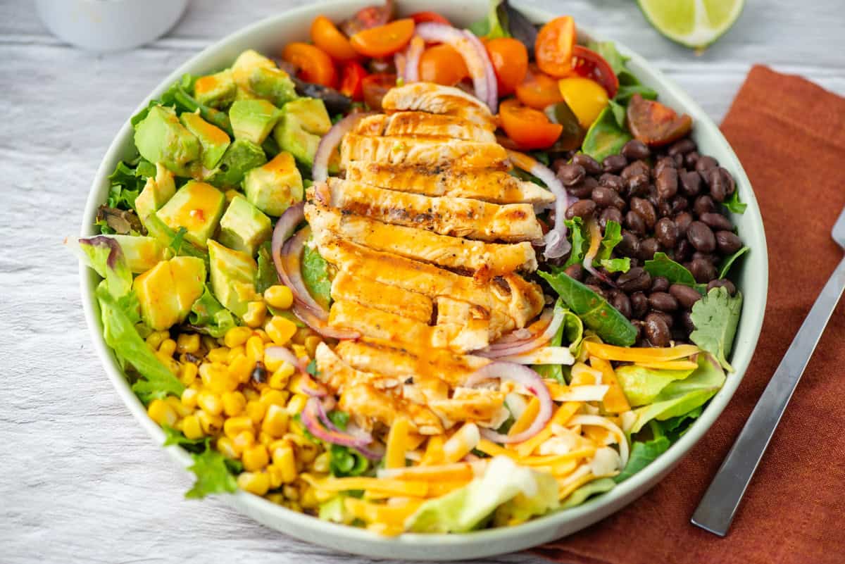 A salad bowl with sliced grilled chicken, black beans, cherry tomatoes, corn, avocado, shredded cheese, red onion, and mixed greens, drizzled with dressing.