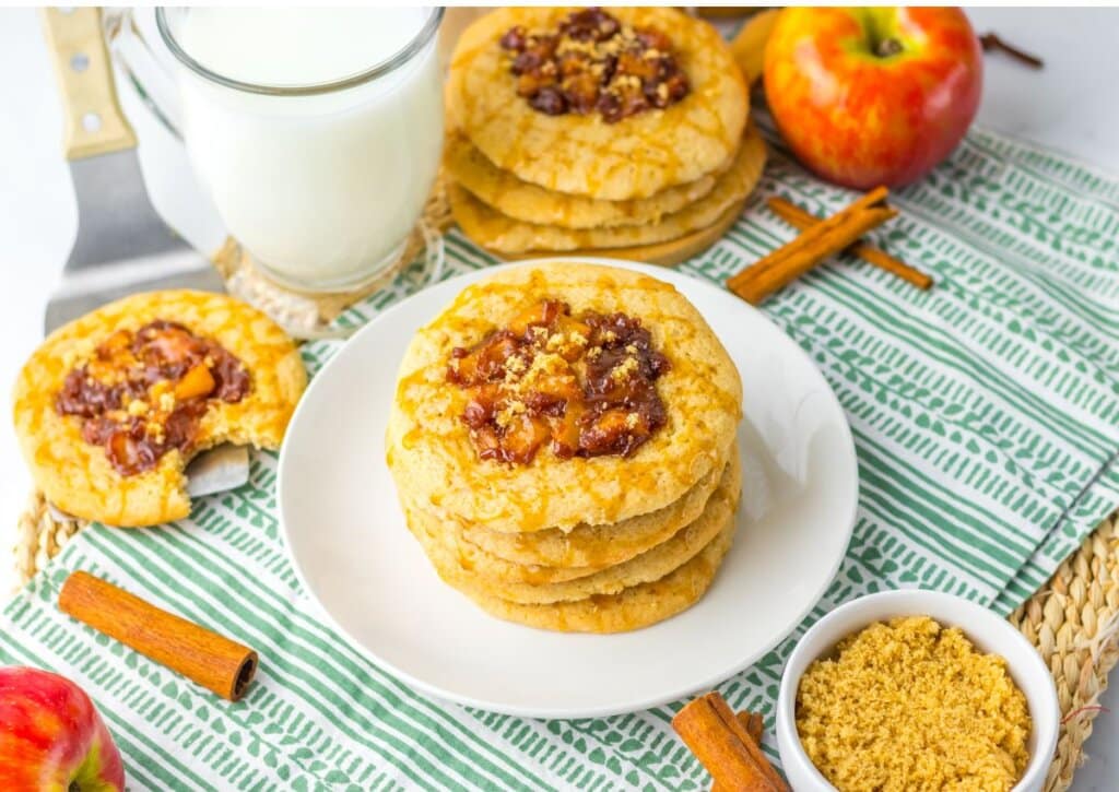 A stack of cookies with fruit topping on a plate, surrounded by apples, cinnamon sticks, brown sugar, and a glass of milk on a striped cloth.