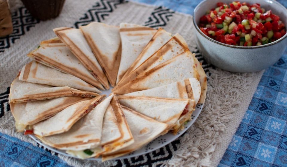 A plate of folded quesadillas arranged in a circle next to a bowl of diced tomato, cucumber, and pepper salsa on a patterned blue tablecloth.