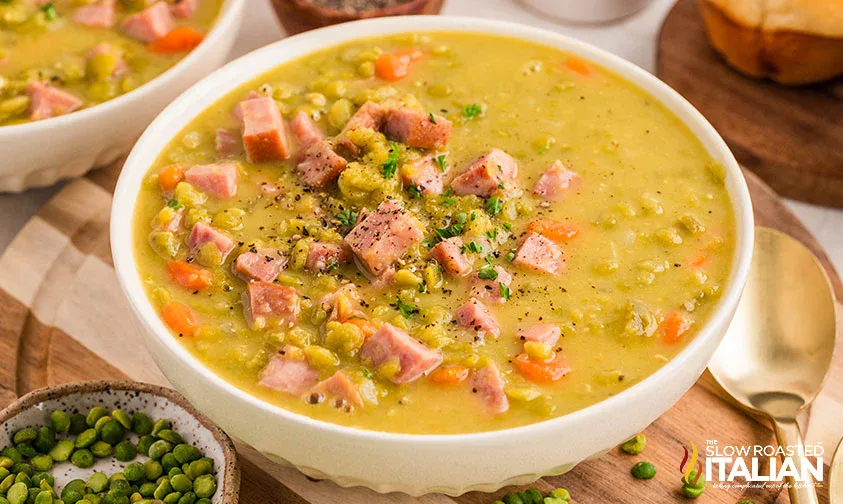 A bowl of split pea soup with diced ham, carrots, and peas, garnished with black pepper, on a wooden surface with a spoon and bread in the background.