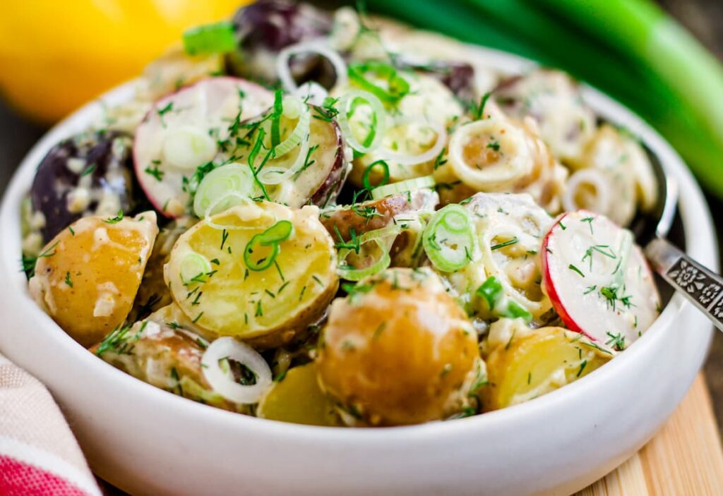 A bowl of potato salad with sliced red and yellow potatoes, radishes, scallions, and fresh herbs, served with a creamy dressing.