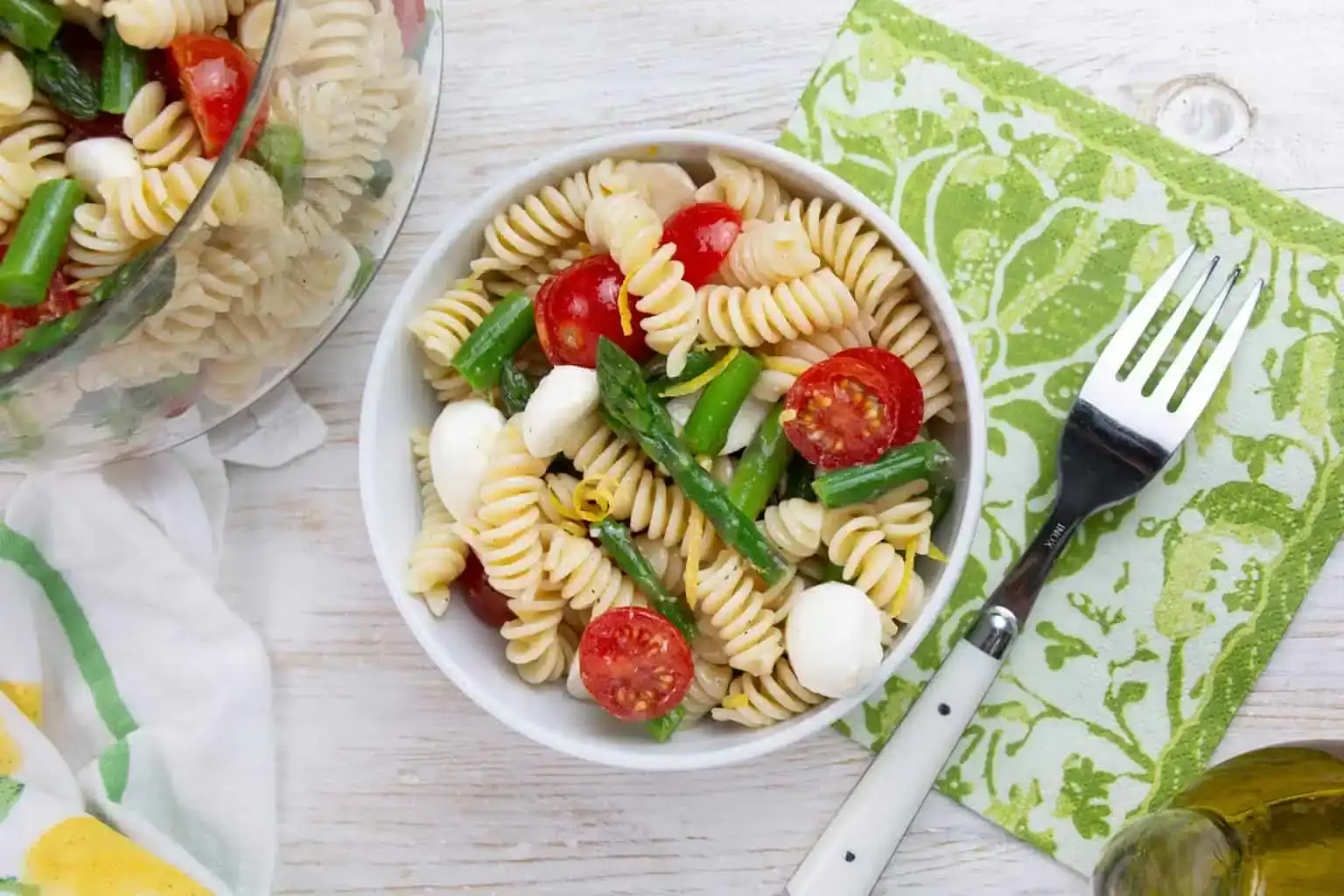 A bowl of pasta salad with rotini, cherry tomatoes, green beans, and mozzarella balls sits on a green napkin beside a fork.
