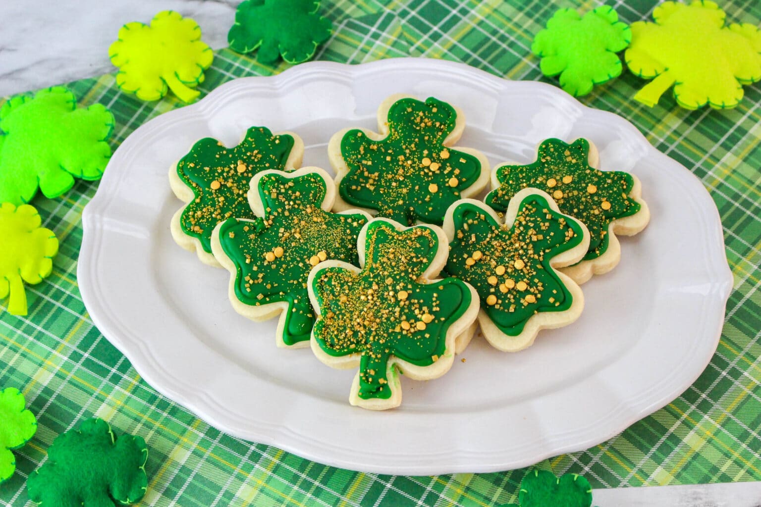 Sugar cookies shaped like shamrocks, decorated with green icing and gold sprinkles, arranged on a white platter with shamrock decorations in the background.
