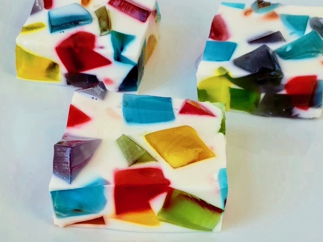 Three square pieces of white gelatin dessert with colorful, translucent jelly cubes embedded inside, displayed on a white surface.