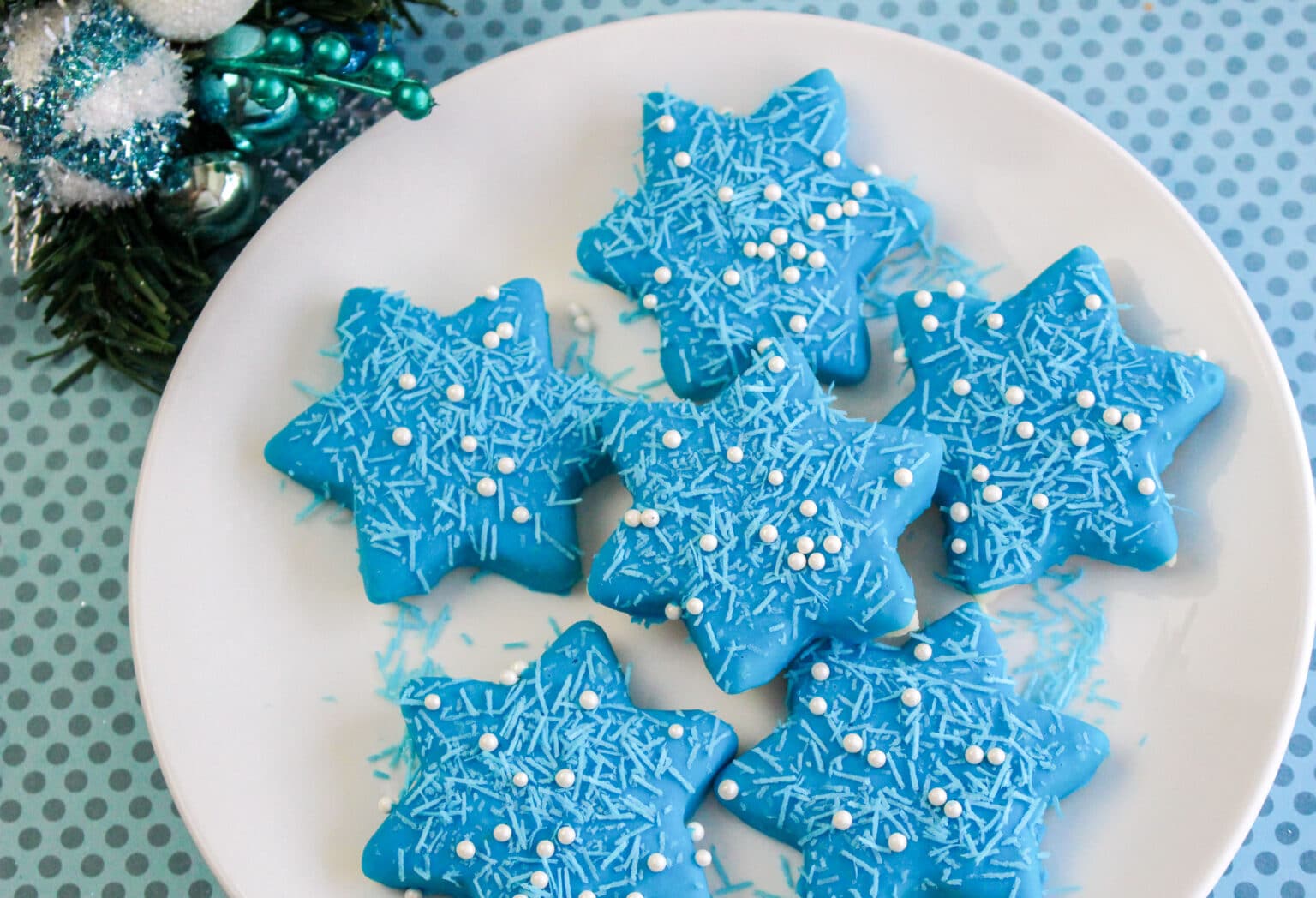 A white plate with six blue star-shaped cookies topped with blue sprinkles and white sugar pearls, placed on a blue polka dot surface.