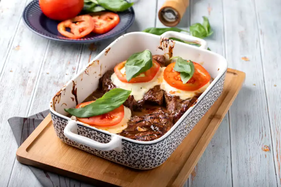 A white baking dish with cooked meat, each piece topped with melted cheese, a tomato slice, and basil leaves, on a wooden board. Sliced tomatoes and basil are in the background.