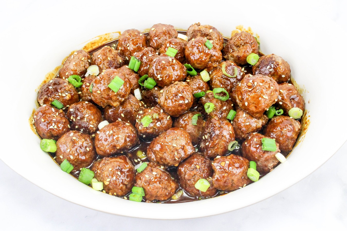 A white bowl filled with glazed meatballs garnished with chopped green onions and sesame seeds.