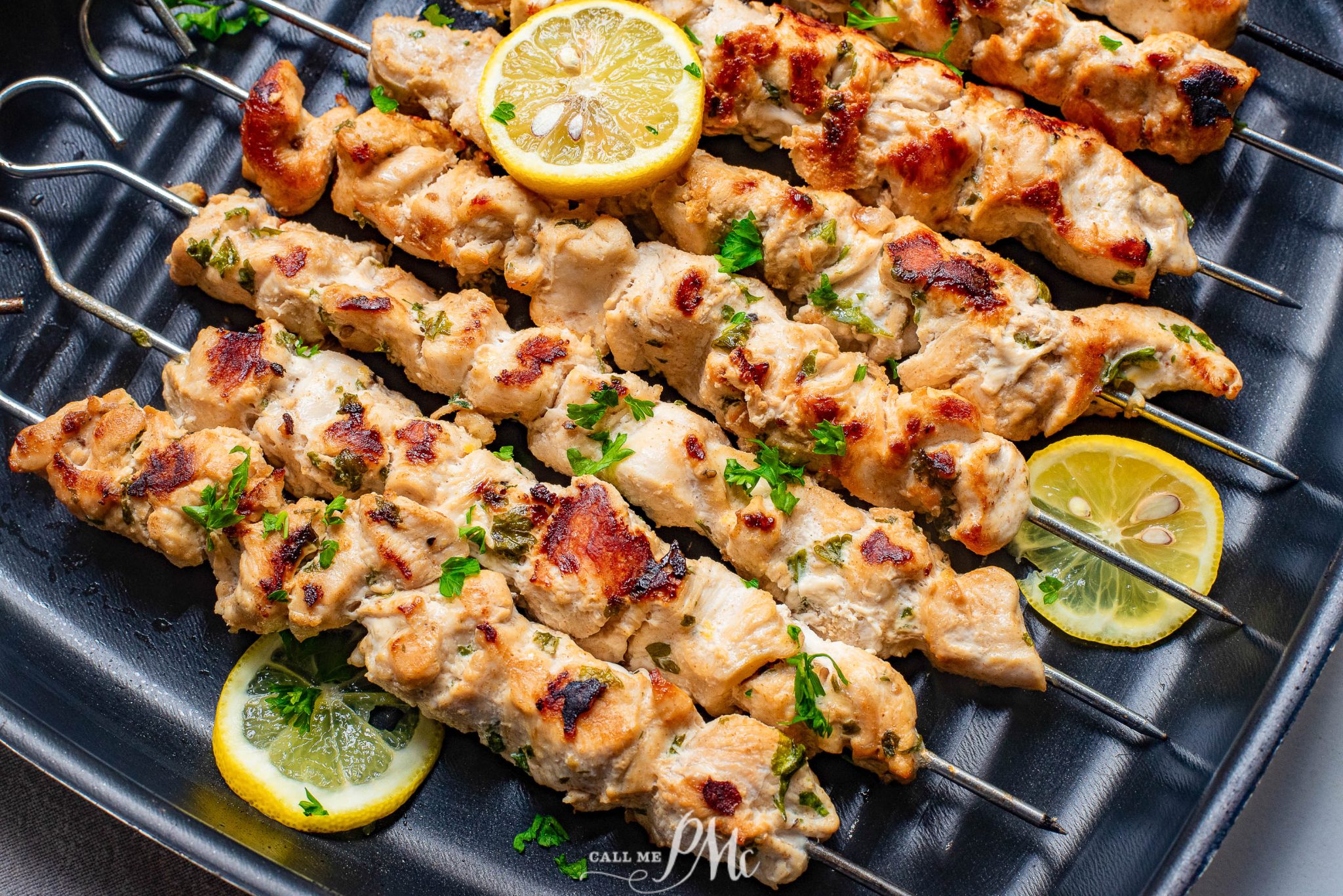 Grilled chicken skewers garnished with parsley and lemon slices, arranged on a ridged black grill pan.
