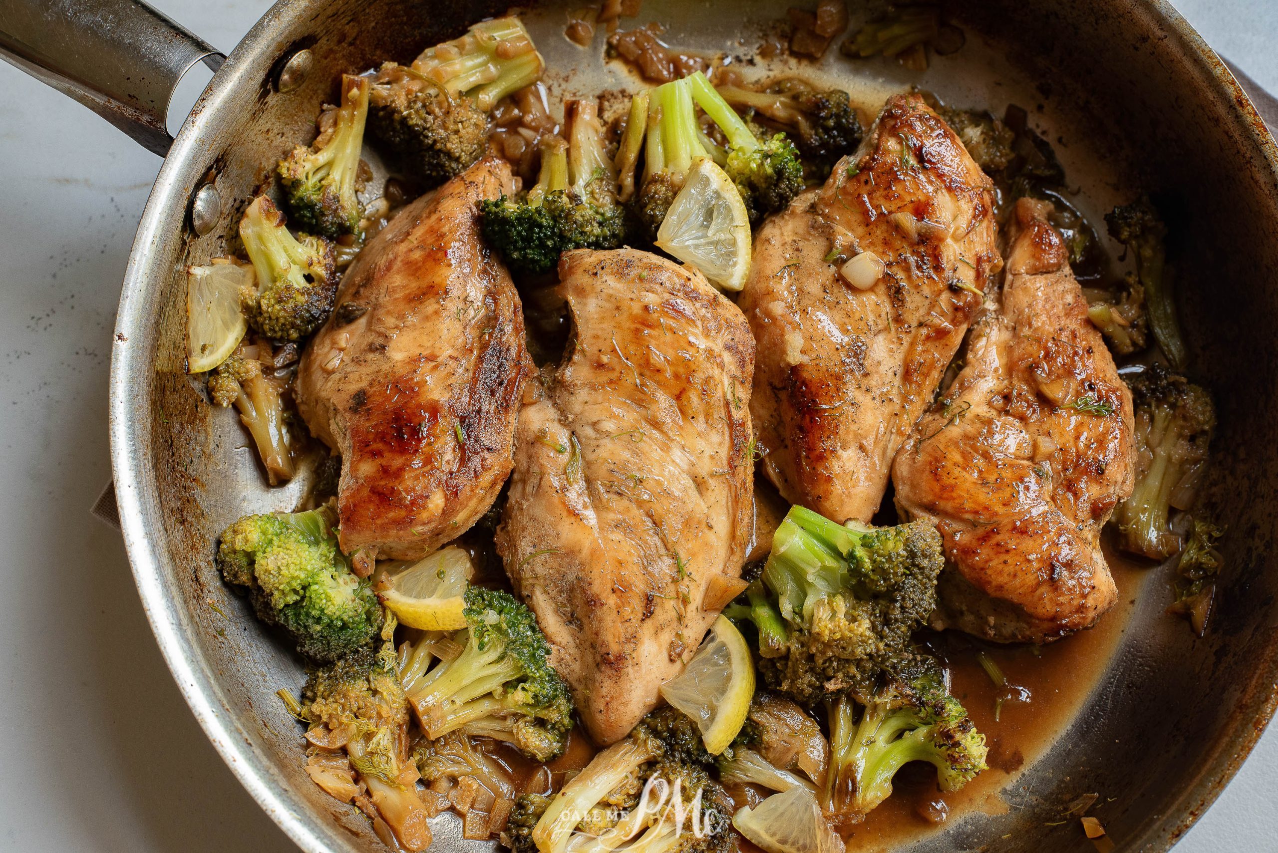 Three cooked chicken breasts with broccoli florets and lemon slices in a skillet with a brown sauce.