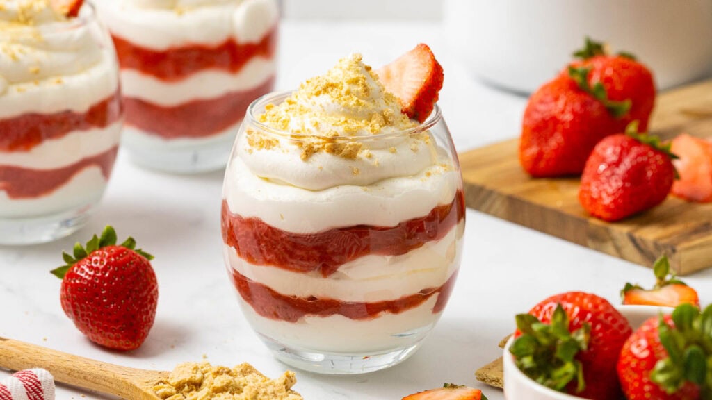 A glass filled with layered whipped cream and strawberry puree, topped with a fresh strawberry and crushed crumbs, with whole strawberries on the side.