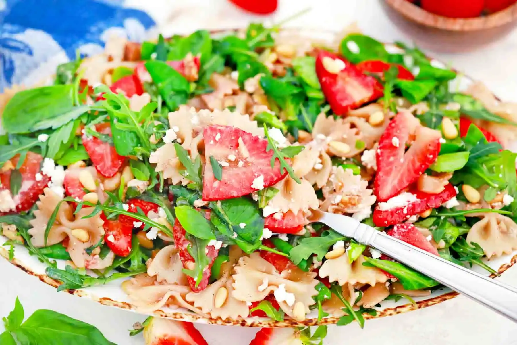 A plate of farfalle pasta salad topped with fresh arugula, sliced strawberries, feta cheese, and pine nuts, with a fork placed on the side.